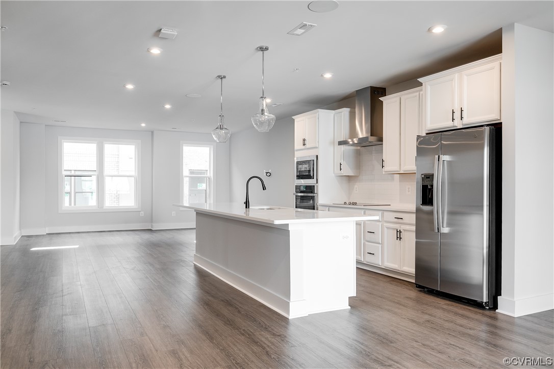 Kitchen with wall chimney range hood, stainless steel appliances, dark hardwood floors, and an island with sink