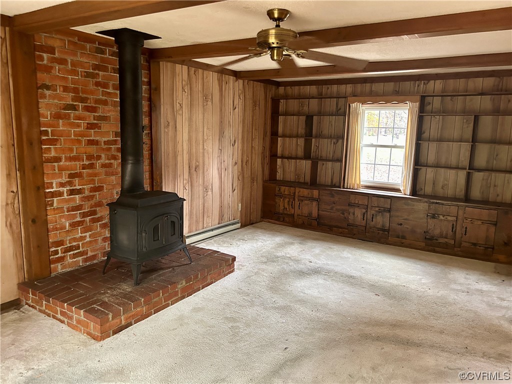 Unfurnished living room featuring light colored carpet, a wood stove, baseboard heating, wood walls, and ceiling fan