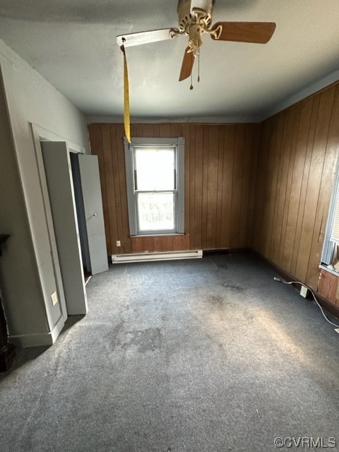Carpeted empty room featuring a baseboard heating unit, ceiling fan, and wooden walls