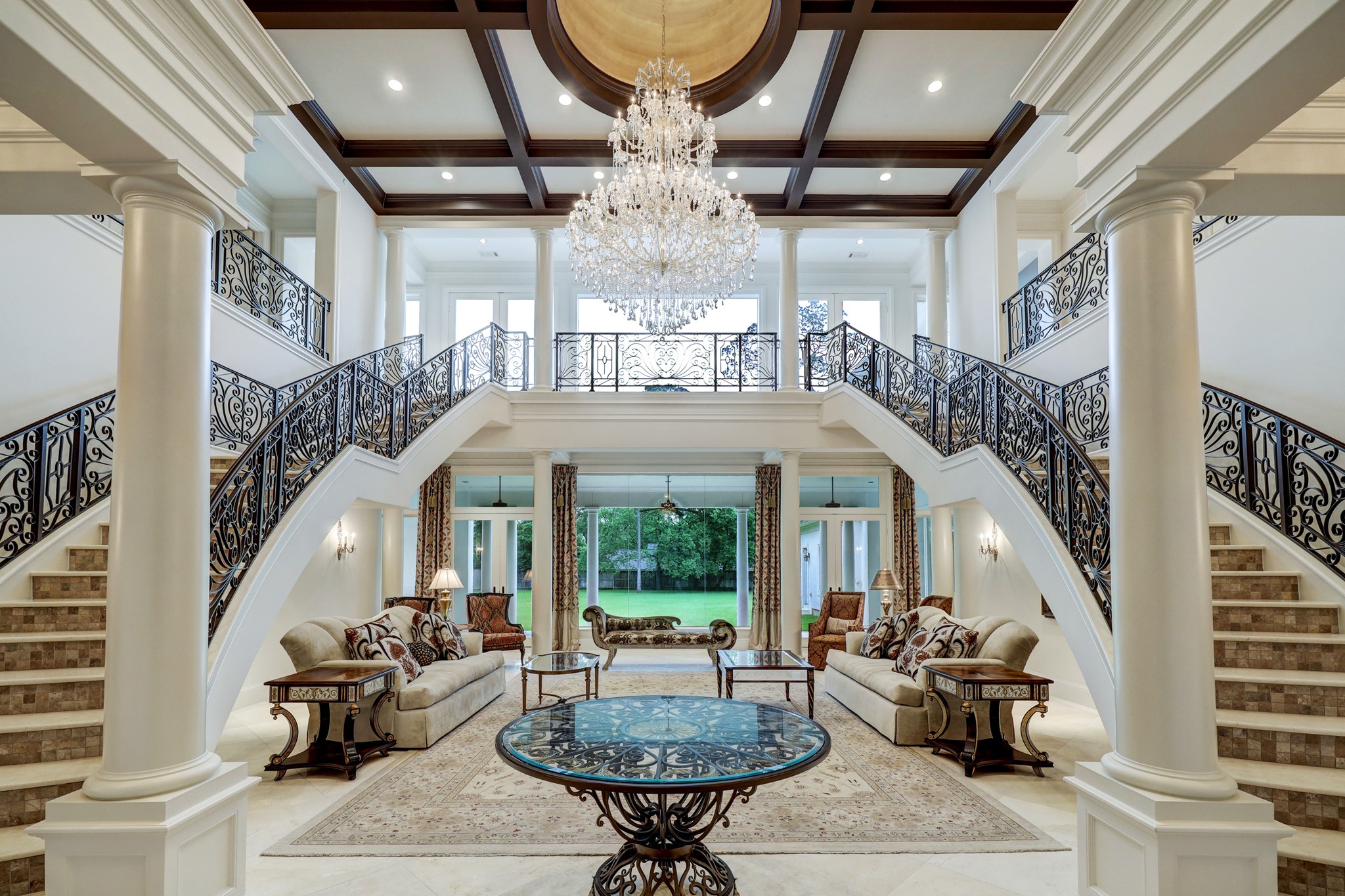The grand two-story foyer opens into the Formal Living room and features double staircases with decorative wrought-iron railings, coffered ceilings, tile floors, decorative columns, a stunning hanging chandelier and great picture window views of the backyard.