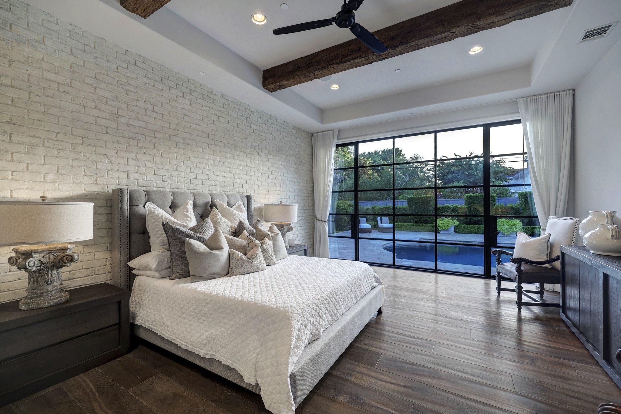 The large primary bedroom includes a painted brick accent wall and a wall of floor-to-ceiling paned windows overlooking your backyard oasis. Exposed wood beams and a trayed ceiling add to the stylish design of the room.