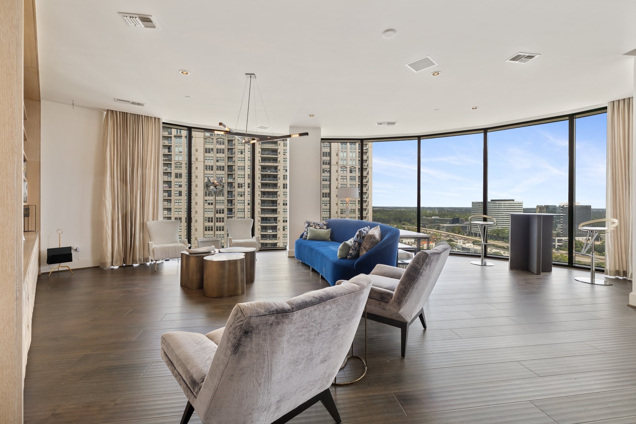 Floor-to-ceiling windows showcasing a city view. Neutral remote shades & curtains hang gracefully on one side, and sleek brand-new flooring adds warmth to the modern space. A minimalist pendant light fixture hangs from the ceiling, completing the elegant ambiance.