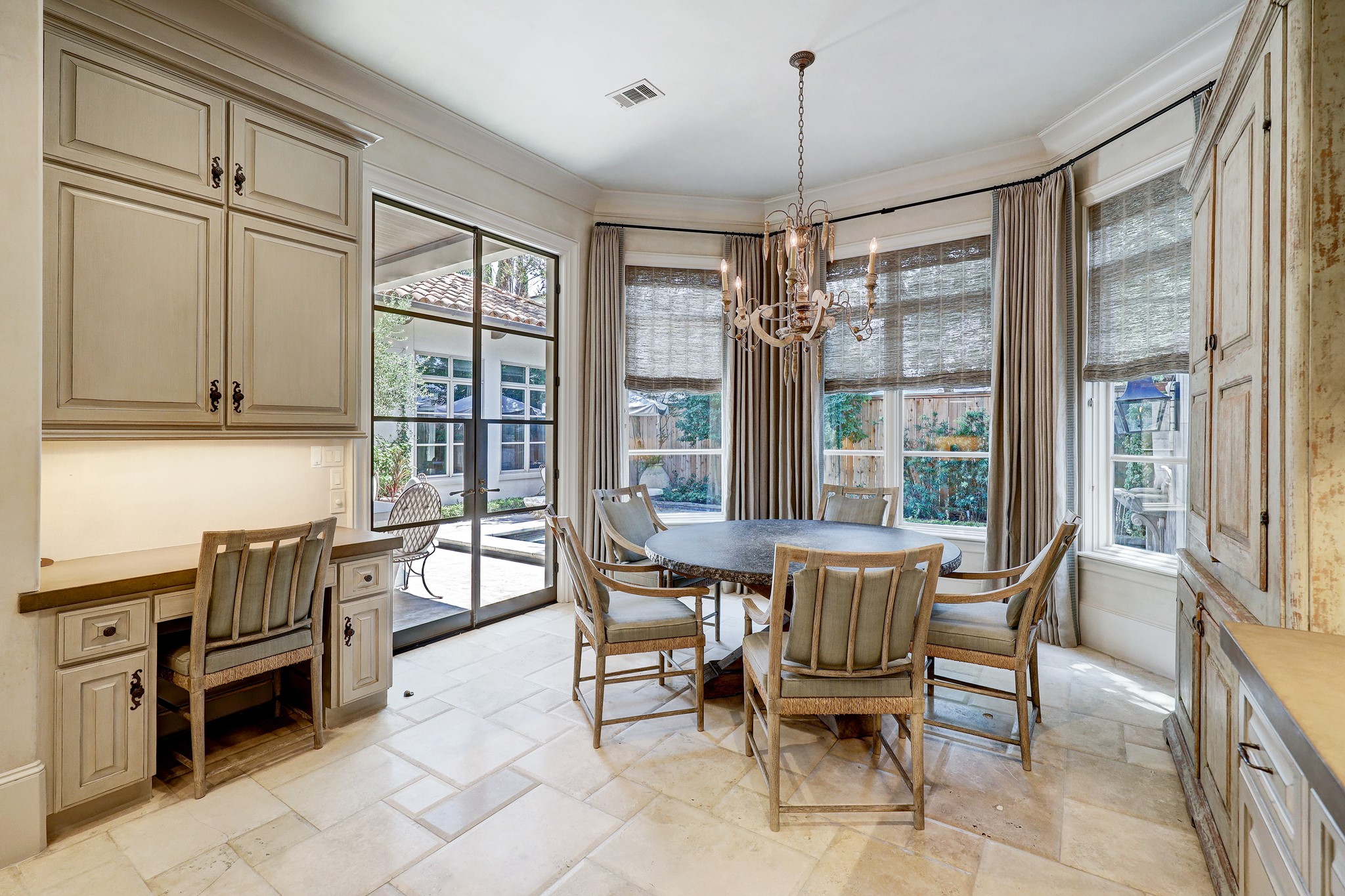 The breakfast area, warmly luminated by a mid-1800s French brass chandelier and the natural light from a vast expanse of windows, offers a panoramic view of the tranquil waters of the pool, while the robust yet elegant steel-framed glass doors provide a stately transition to the veranda.