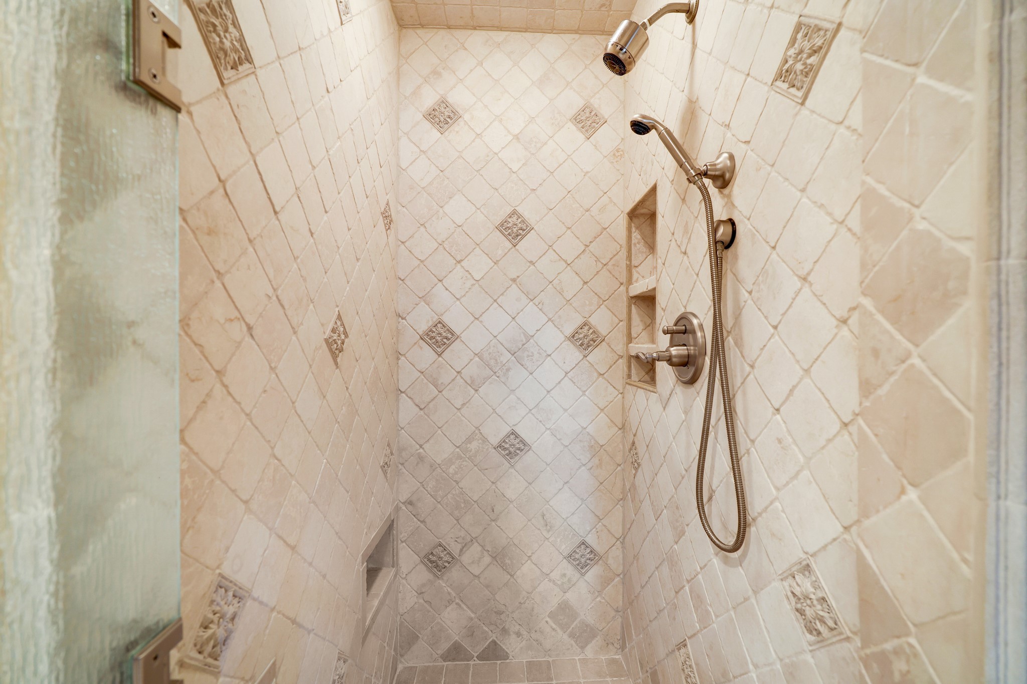 The shower is designed with a diamond-patterned Italian tile, accented with elegant decorative insets, offering a timeless look that complements the serene palette of the room. Modern fixtures, including a versatile handheld showerhead, ensure a spa-like experience in the comfort of your own home.