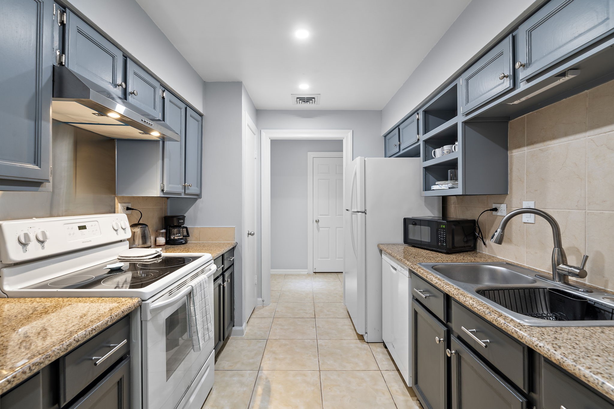 Galley kitchen with gray-blue cabinets, stone countertops, leading to a doorway.