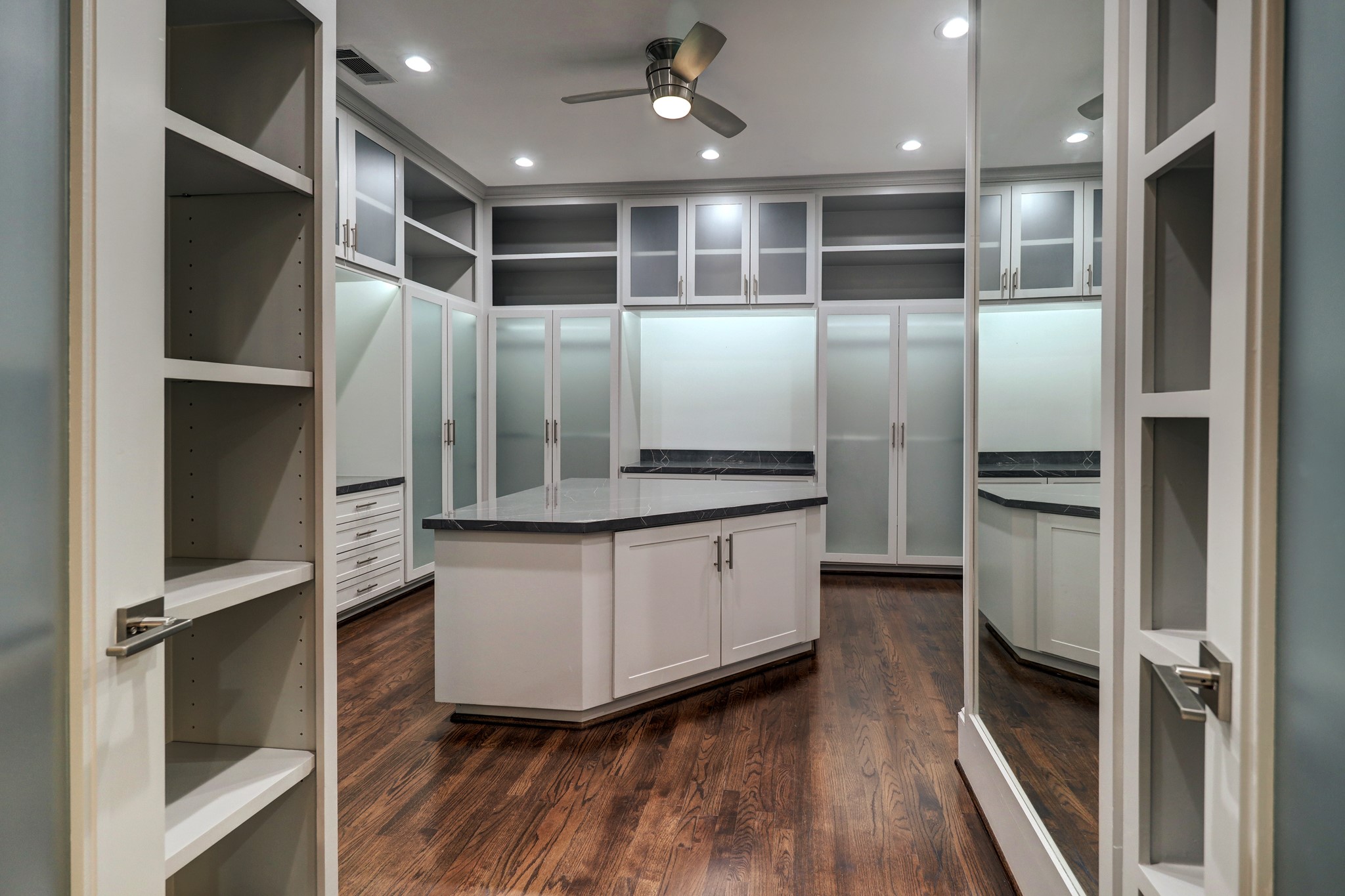The Primary Walk-in Closet is huge and features hardwood floors, built-in floor length mirror, tons of cabinets (some of which are frosted glass-faced) and drawers for clothes and accessories of all sizes and types, and a large packing island with additional storage.