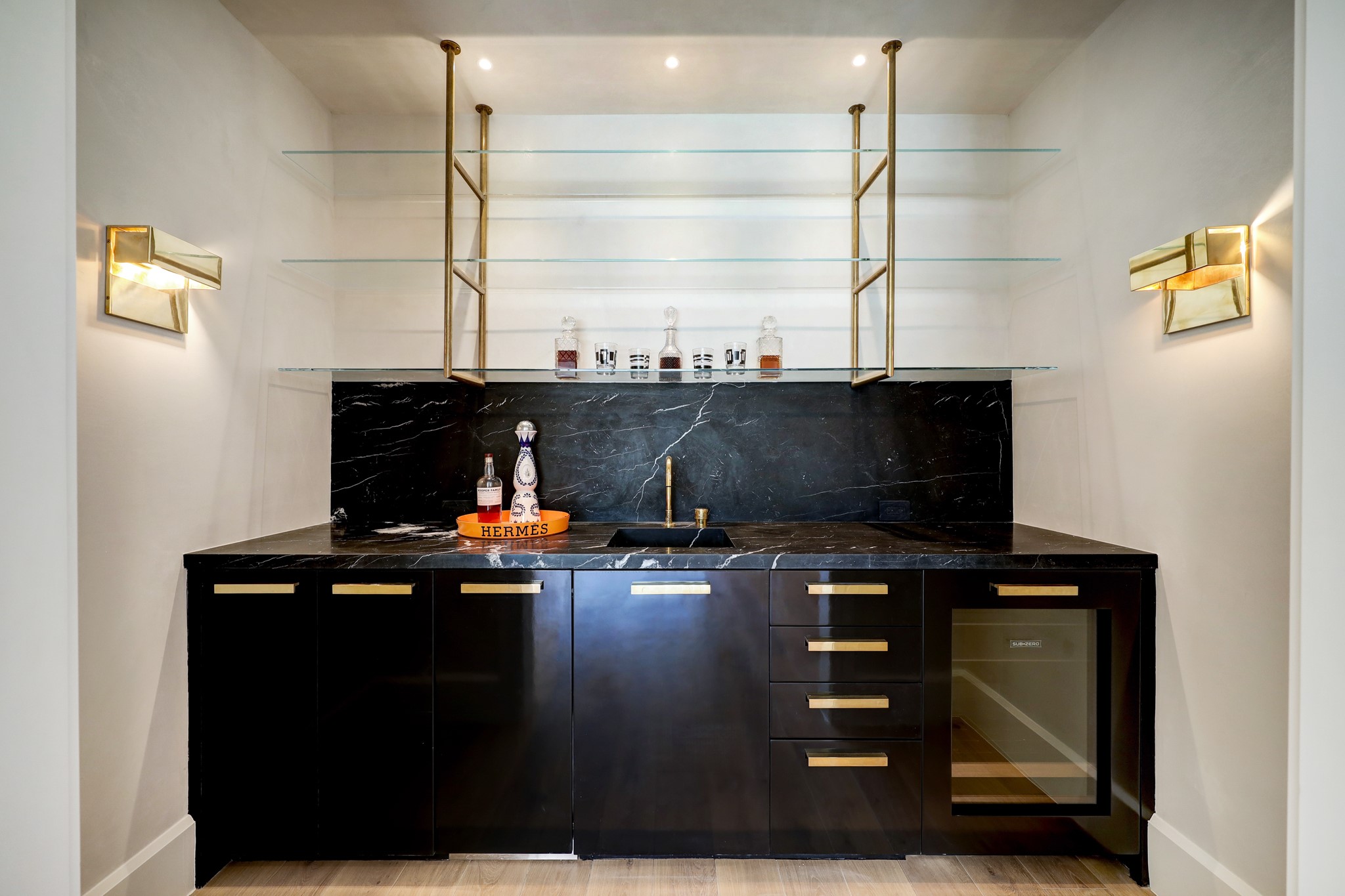 An intimate view of the wet bar, finished in high-gloss lacquered paint and adorned with unlacquered brass hardware. The sunken sink, crafted in marble, adds an elegant touch. The space is fully equipped with an under-mounted fridge, ample storage, and an ice maker.