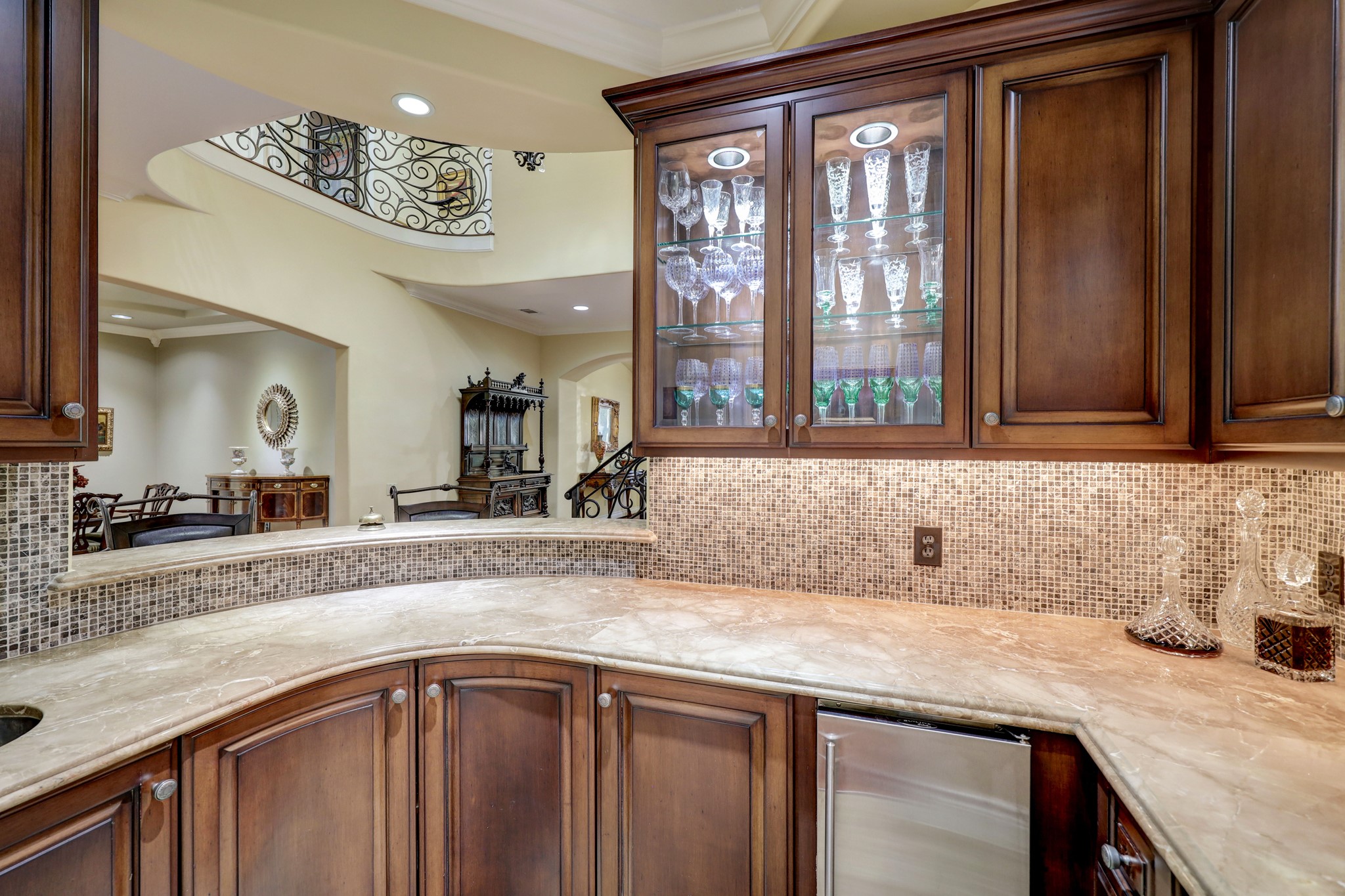 The Walk-in Wet Bar [9 x 6] features marble countertops with a mosaic tile backsplash, round bar sink, icemaker, under cabinet lighting, glass front display cabinets with glass shelves and lighting, and a pass through to the Foyer.