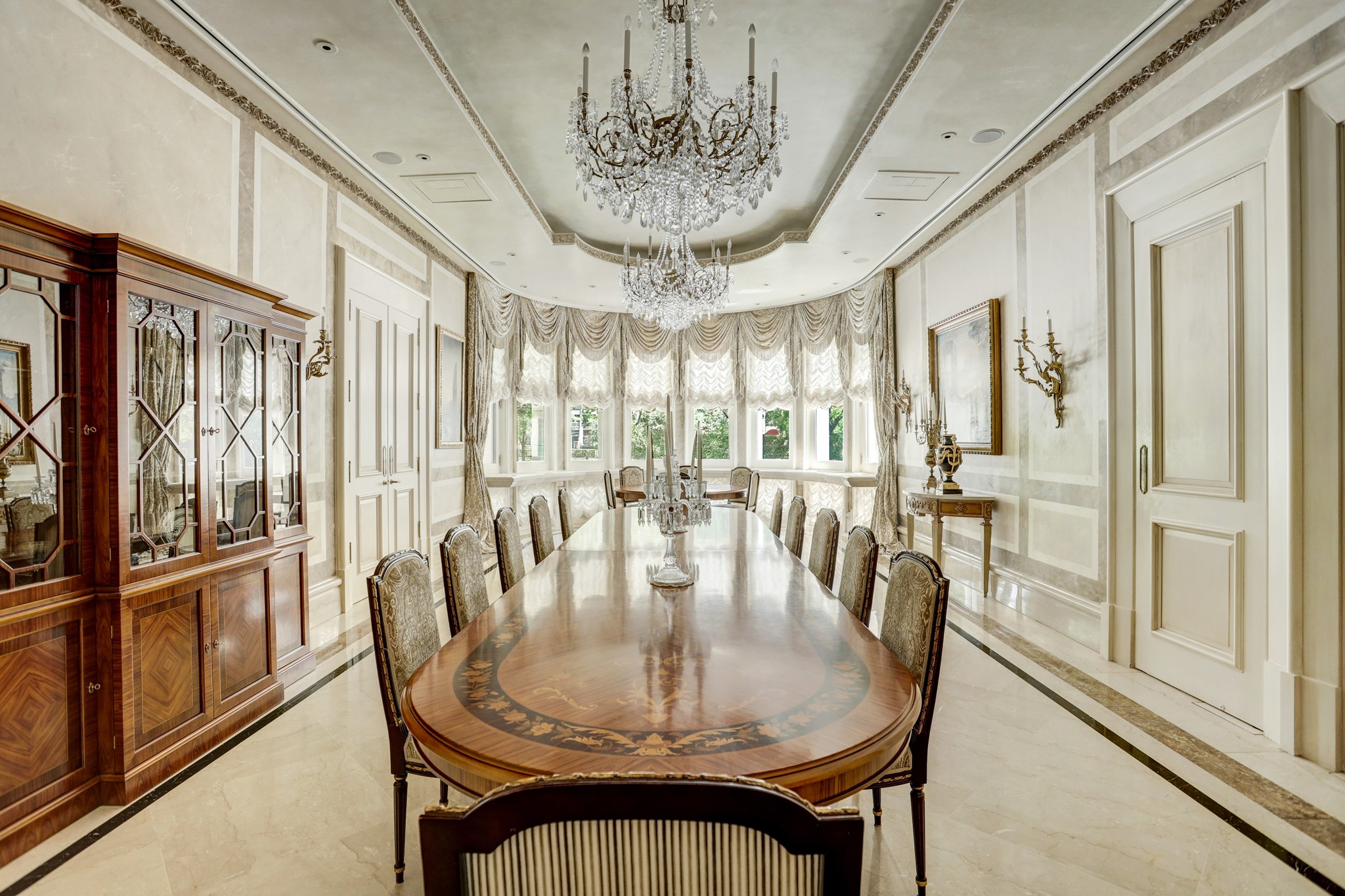 The grandiose formal dining room seats 44 and features an expertly-crafted wall finish which perfectly matches the sleek, inlaid marble tile floor.  An ornamental plastered ceiling boasts recessed cove lighting while natural light floods the space through the extensive bow window which features custom, multi-layered balloon valances. To the left of the frame, notice the swinging double doors which offer easy access to the Butler’s pantry and climate-controlled wine vault. 
