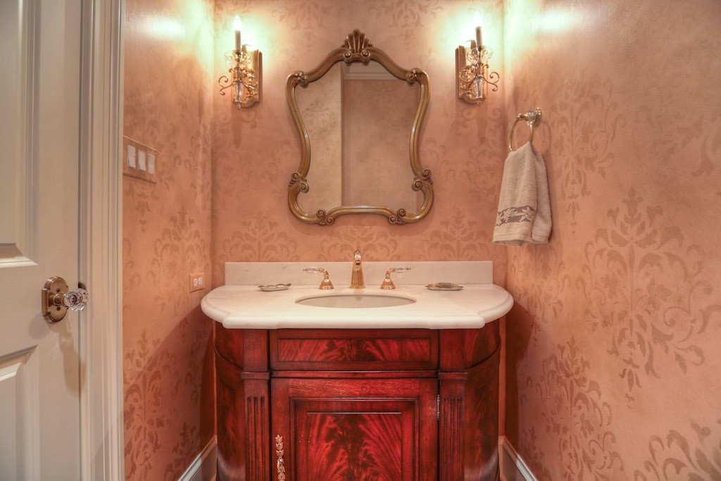 Powder bath has custom stained Crotch Mahogany sink console with Sugar White Marble countertop with gold fixtures. Hand-painted stenciled walls in a custom design. A pair of Waterford crystal sconces flank the mirror.