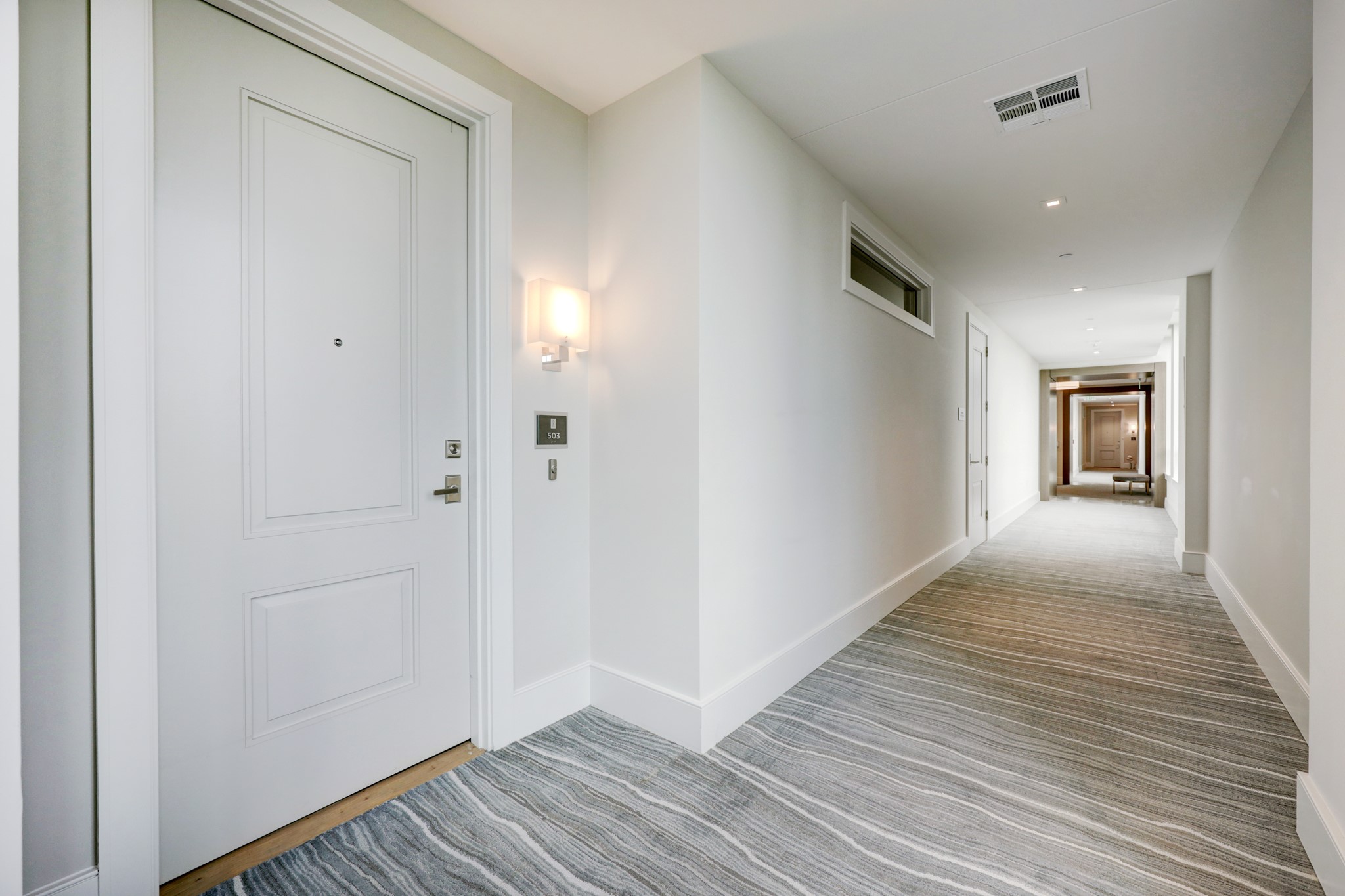View of the FRONT DOOR to #503. A brightly lit hallway from the elevator shaft leads through the softly painted walls and handsome carpeting to the unit's stately front door with elegant door handle/wall sconce.