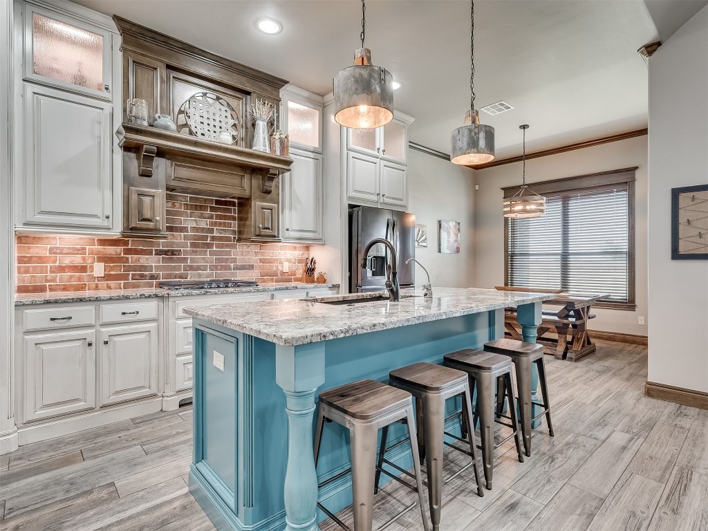 1123 S Czech Hall Road, Tuttle, OK 73089 kitchen featuring appliances with stainless steel finishes, a center island, white cabinets, light stone countertops, hanging light fixtures, light hardwood flooring, kitchen island with sink, ornamental molding, and backsplash
